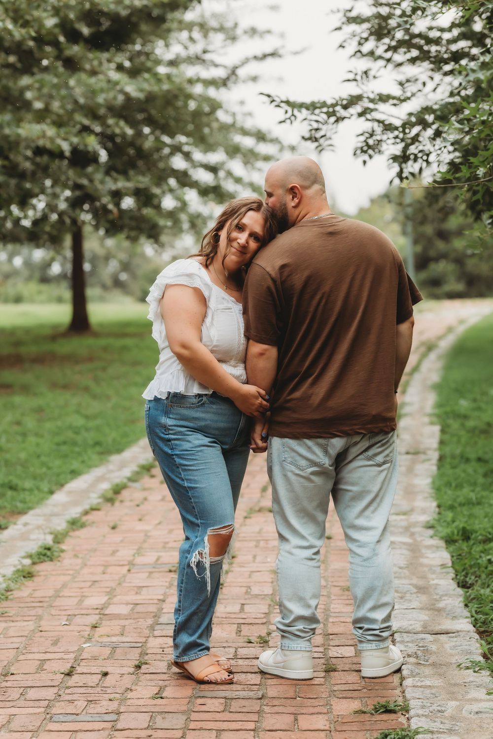Couple holding hands on a brick path in a lush green park, woman in white top and jeans leaning on man.