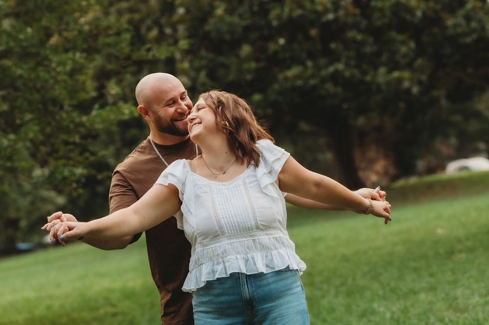 A happy couple dances outdoors in a lush green park, the woman smiling with arms outstretched in a joyful embrace.