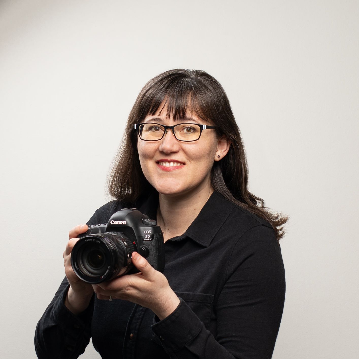 Headshot portrait of Holli wearing a black shirt and black glasses smiling at camera with a white background, color photo.