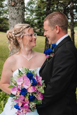 Bride and groom share intimate moment outdoors with pink and blue bouquet and natural backdrop.