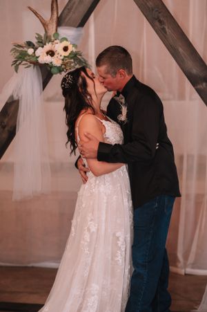 A romantic wedding kiss under a wooden arch decorated with white flowers and sheer curtains.