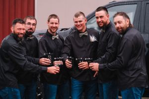Groomsmen in black shirts and jeans posing with beverages for a casual wedding photo.