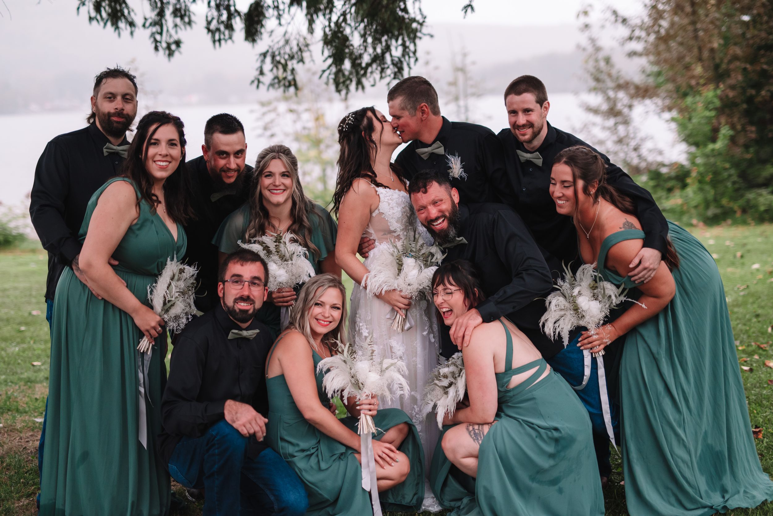 Wedding party in green dresses and black shirts pose playfully by a lake.