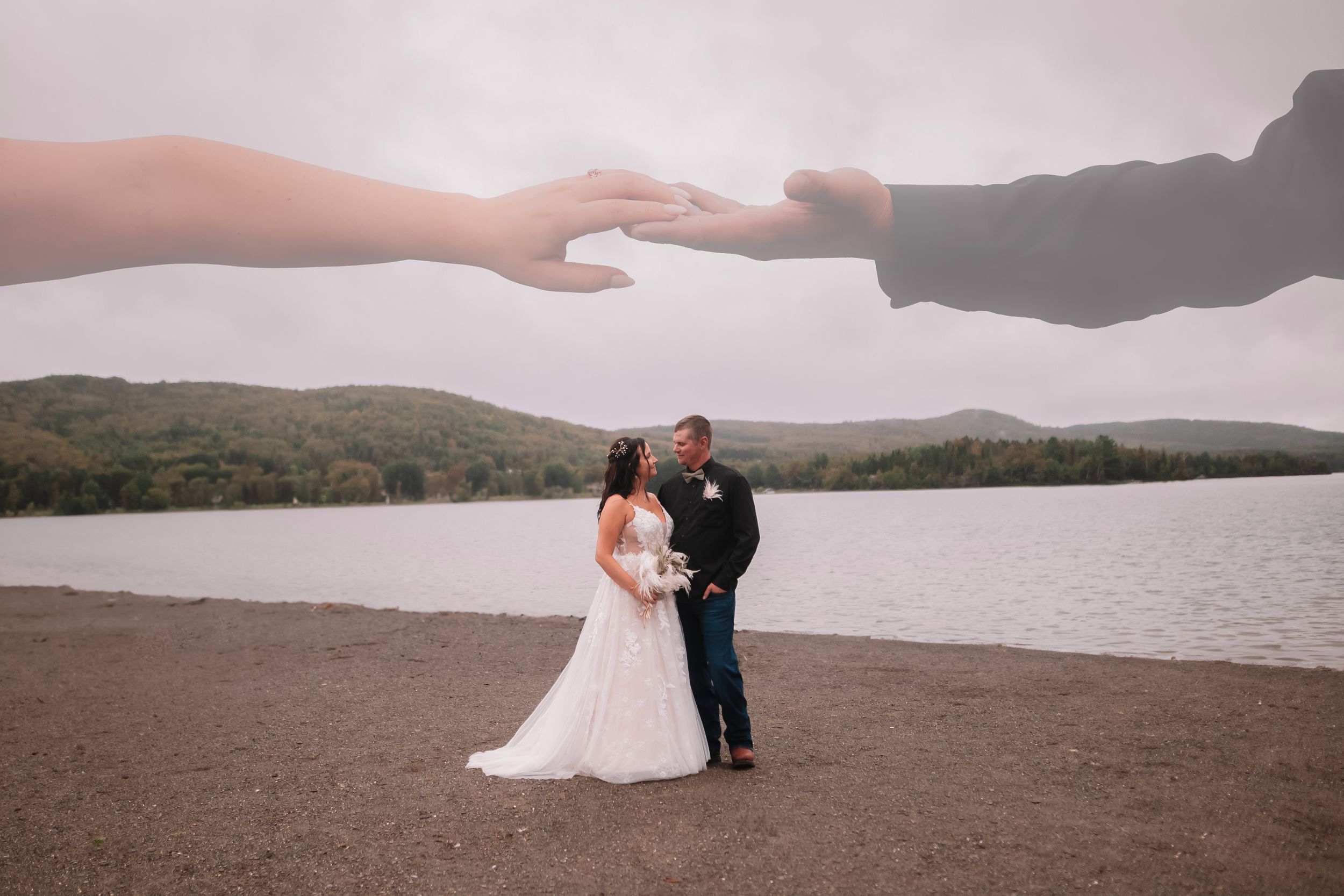 Creative wedding photo with hands reaching across the sky above a couple by a lake.