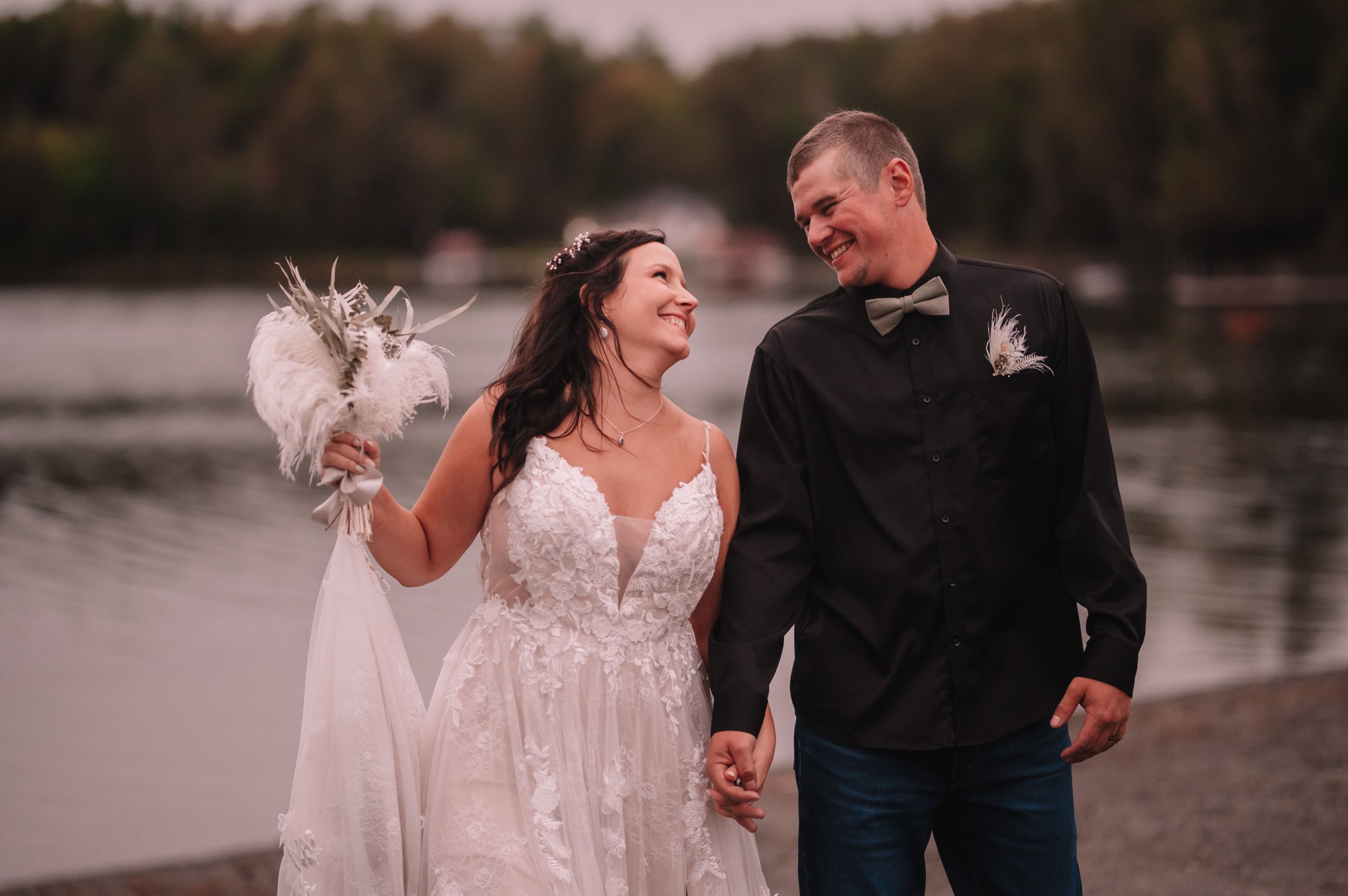 Joyful newlyweds walking hand in hand by a serene lake at sunset.