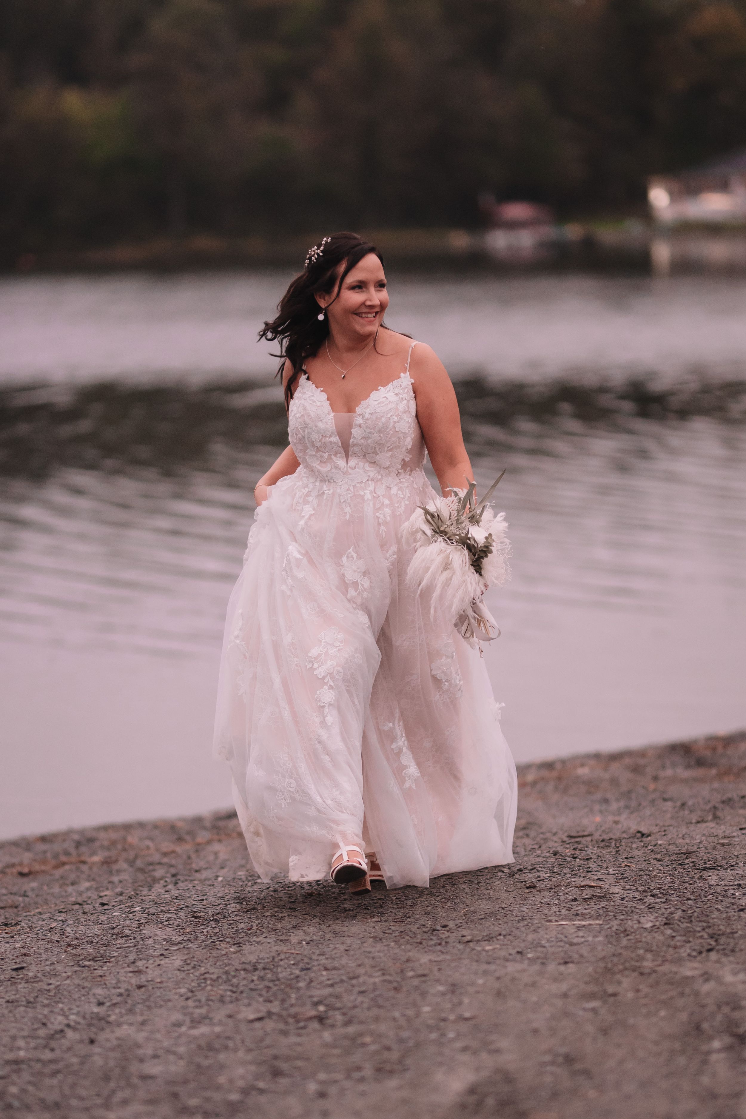 Bride in a flowing lace wedding gown walks along a lakeside shore holding a dried floral bouquet at dusk.