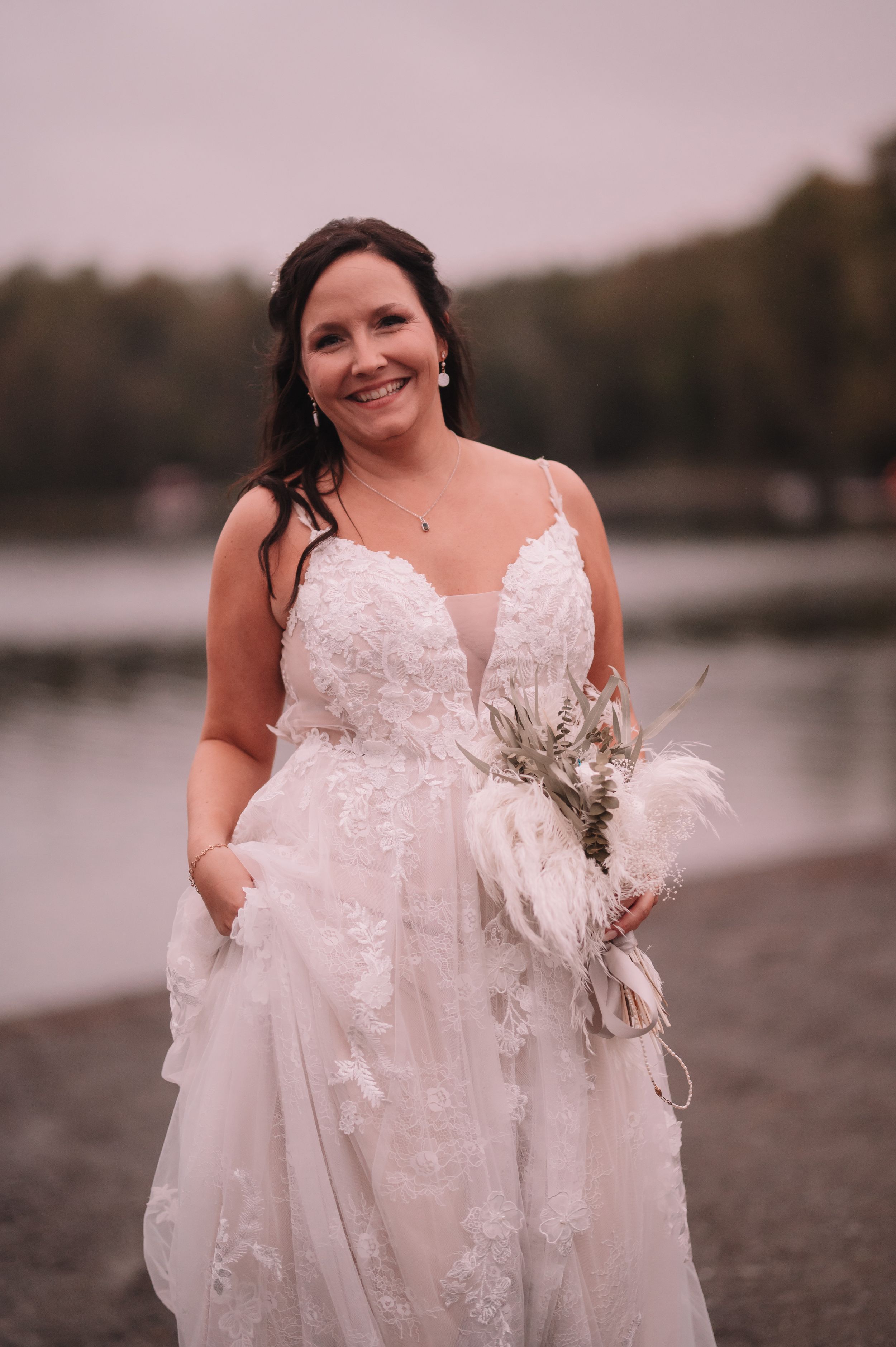 Elegant bride in a detailed lace wedding dress poses by a lake with a rustic bouquet.