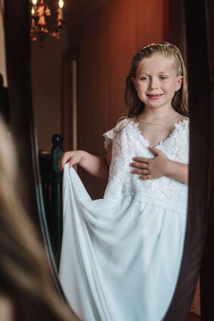 Young flower girl in white lace dress smiling while looking at her reflection.