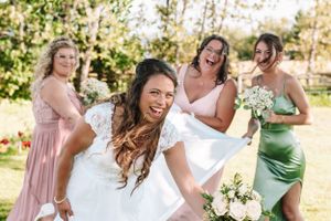 Candid moment of bride and bridesmaids laughing and dancing outdoors in pastel dresses.