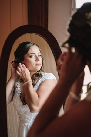 Bride putting on earrings while looking in oval mirror during wedding preparations.