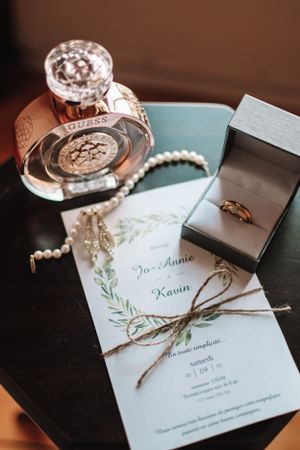 Wedding details flatlay with invitation, pearl necklace, perfume bottle and engagement ring in box.