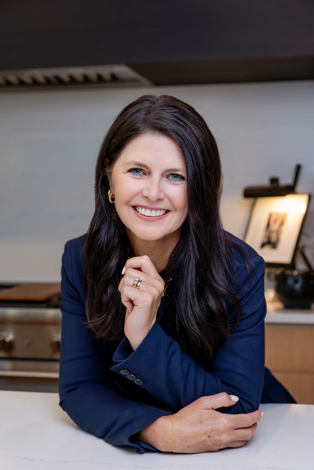 Professional headshot of someone in a navy blazer smiling warmly while leaning on a white desk in an office setting.