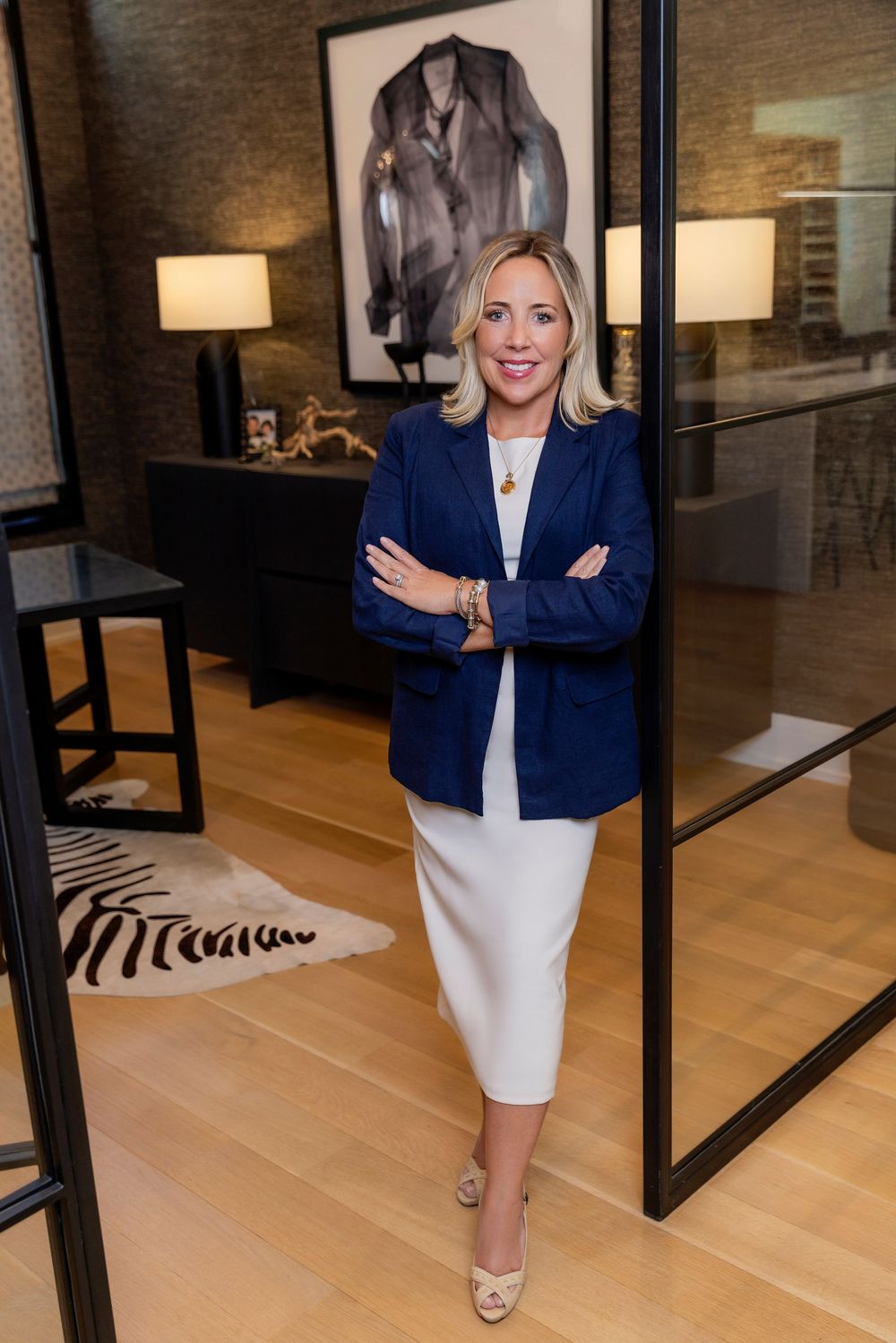 Professional wearing a navy blazer and white dress stands confidently in a modern office with artwork and decor.