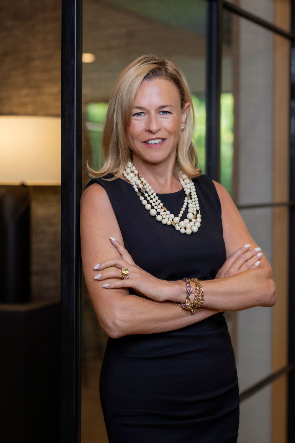 Professional business portrait of someone in a black sleeveless dress with pearl necklace and bracelets in an office setting.