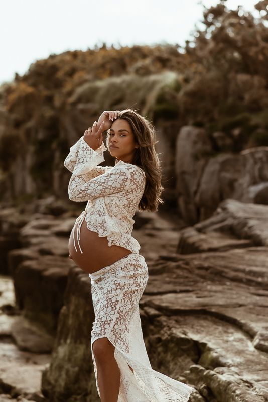 A model in a white lace crop top poses against a rocky beach landscape in warm sepia tones.