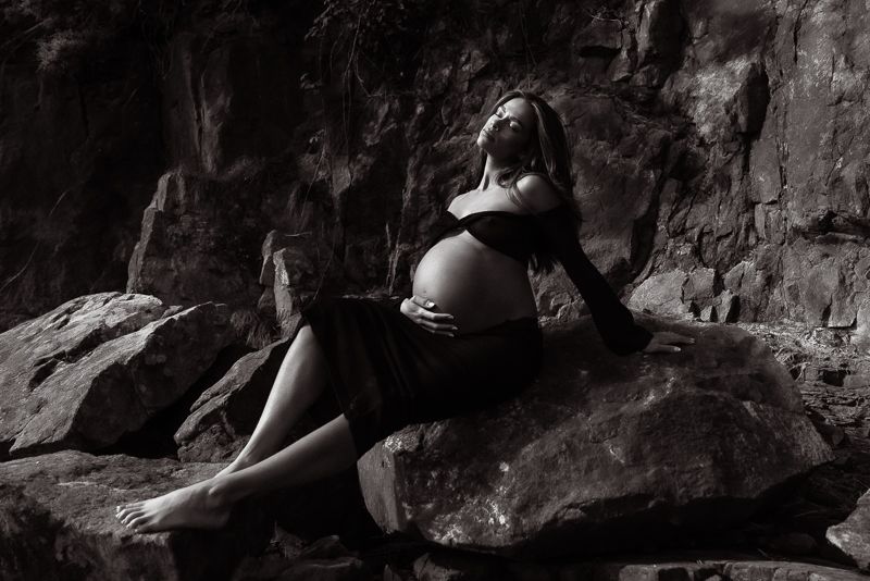 A dramatic black and white maternity portrait shows an expectant mother posing on rocky terrain.