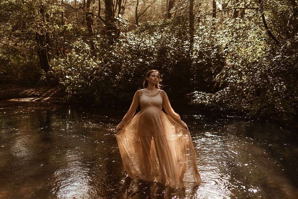A person in a flowing champagne-colored gown stands gracefully in a misty forest stream during golden hour In Chorley woodland