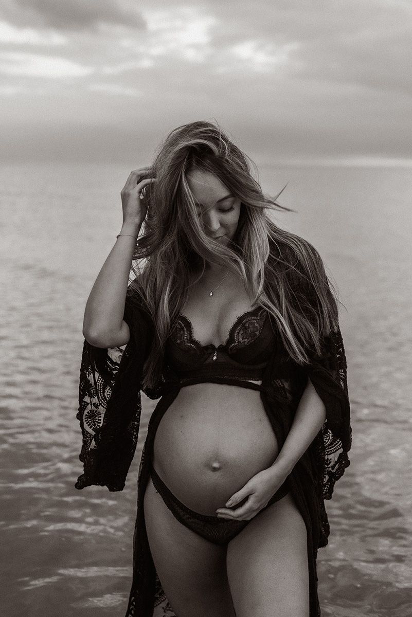 Black and white maternity photo showing pregnant woman in lace lingerie standing by ocean at sunset.