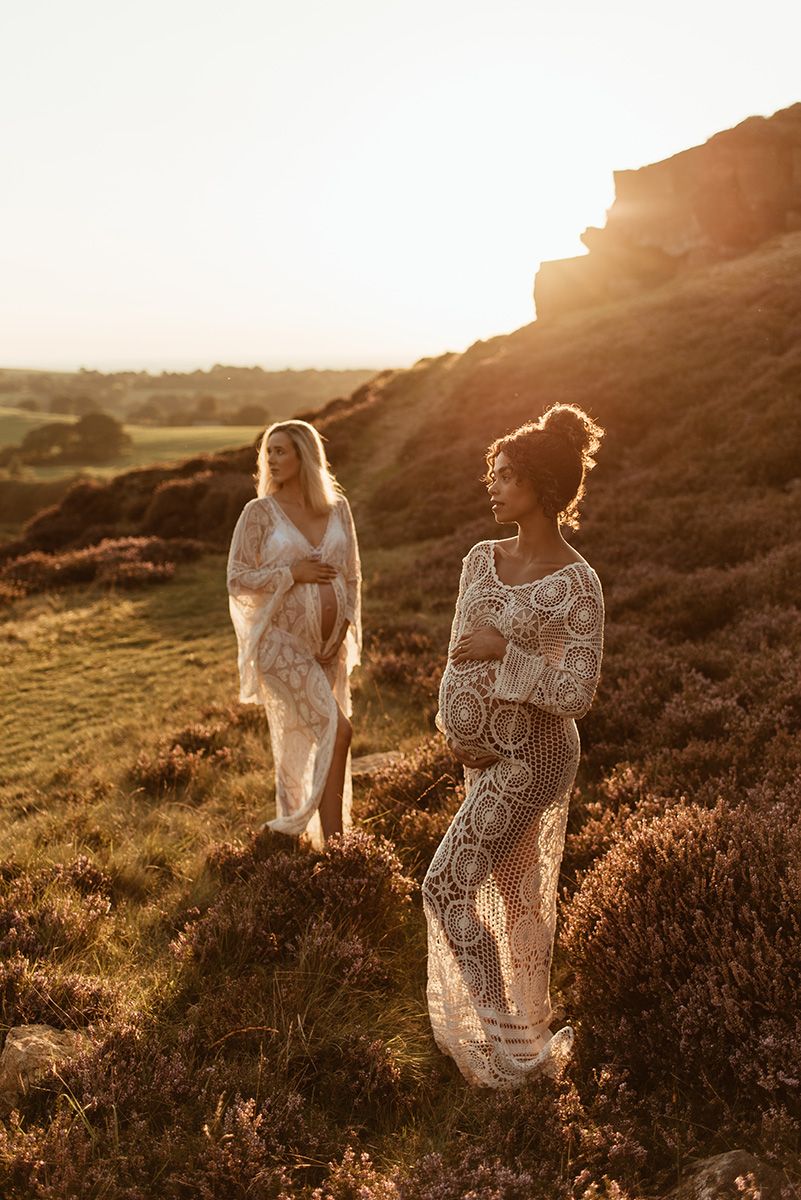Two pregnant women in flowing lace dresses stand on a grassy hillside during golden hour sunset maternity Photoshoot