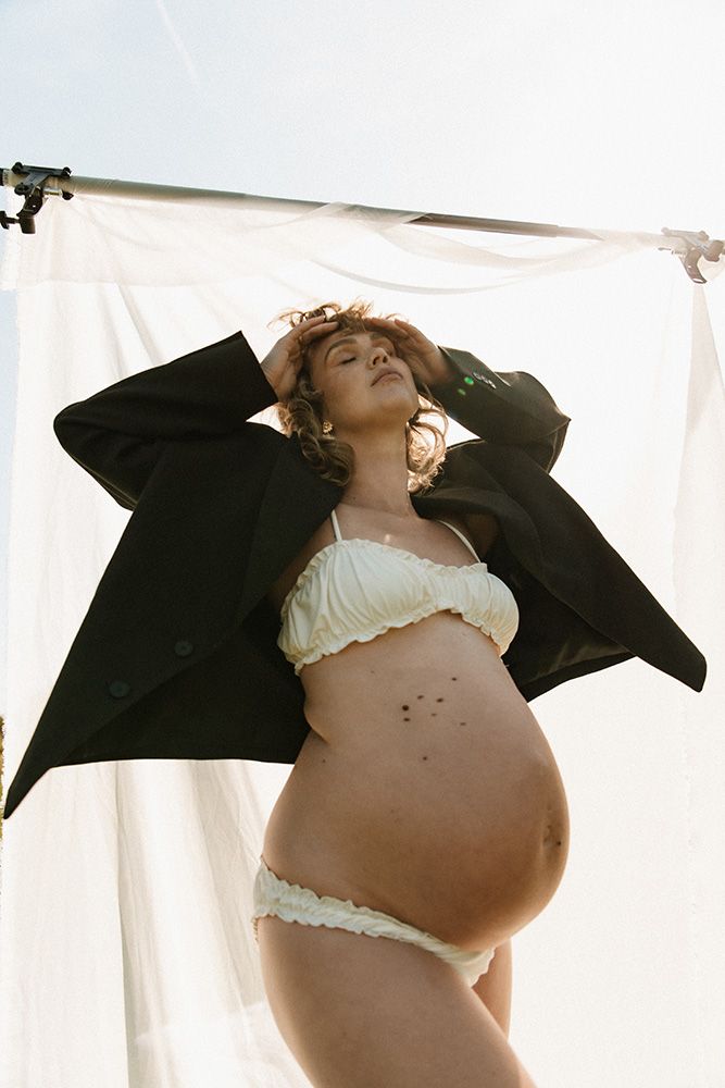 Maternity photo of pregnant woman in white lingerie and black jacket posing against white backdrop outdoors.