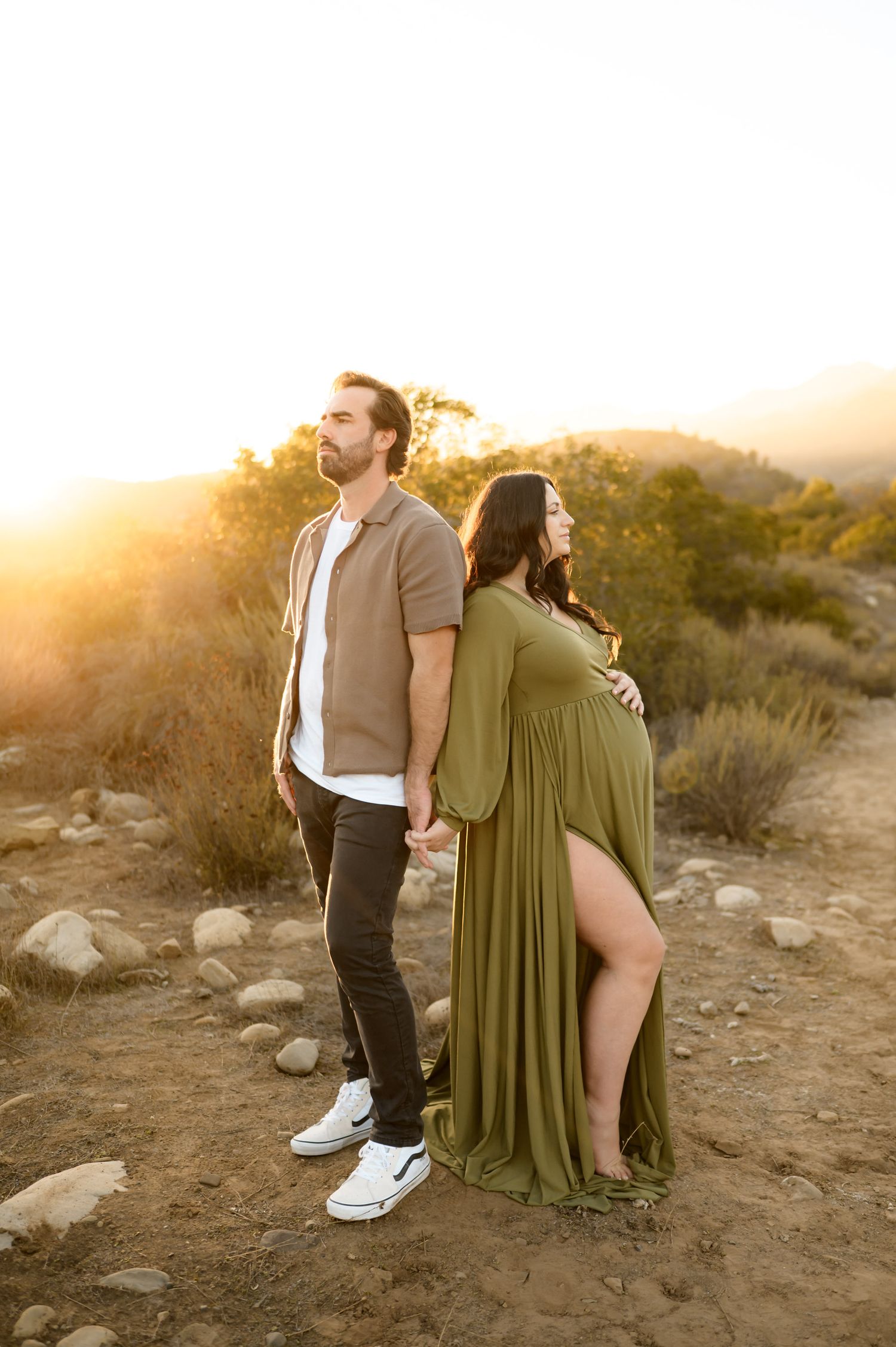 couple stands back to back at sunset Ojai maternity photoshoot. Woman wears long green dress. By Ventura, Santa Barbara photographer