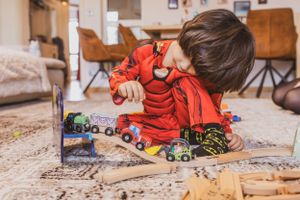 Foto einer Familien Homestory, das ein Kind im Superheldenkostüm zeigt, das auf dem Teppich sitzt und mit Schienen und Fahrzeugen spielt.