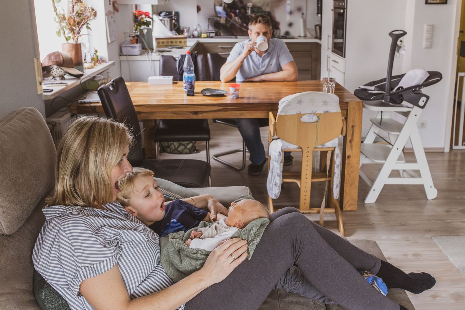 Mutter mit Neugeborenem und großem Bruder auf dem Sofa, das Baby gähnt, die beiden ahmen es nach und im Hintergrund trinkt der Vater Kaffee.