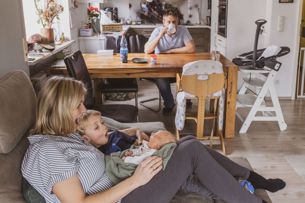 Mutter mit Neugeborenem und großem Bruder auf dem Sofa, das Baby gähnt, die beiden ahmen es nach und im Hintergrund trinkt der Vater Kaffee.