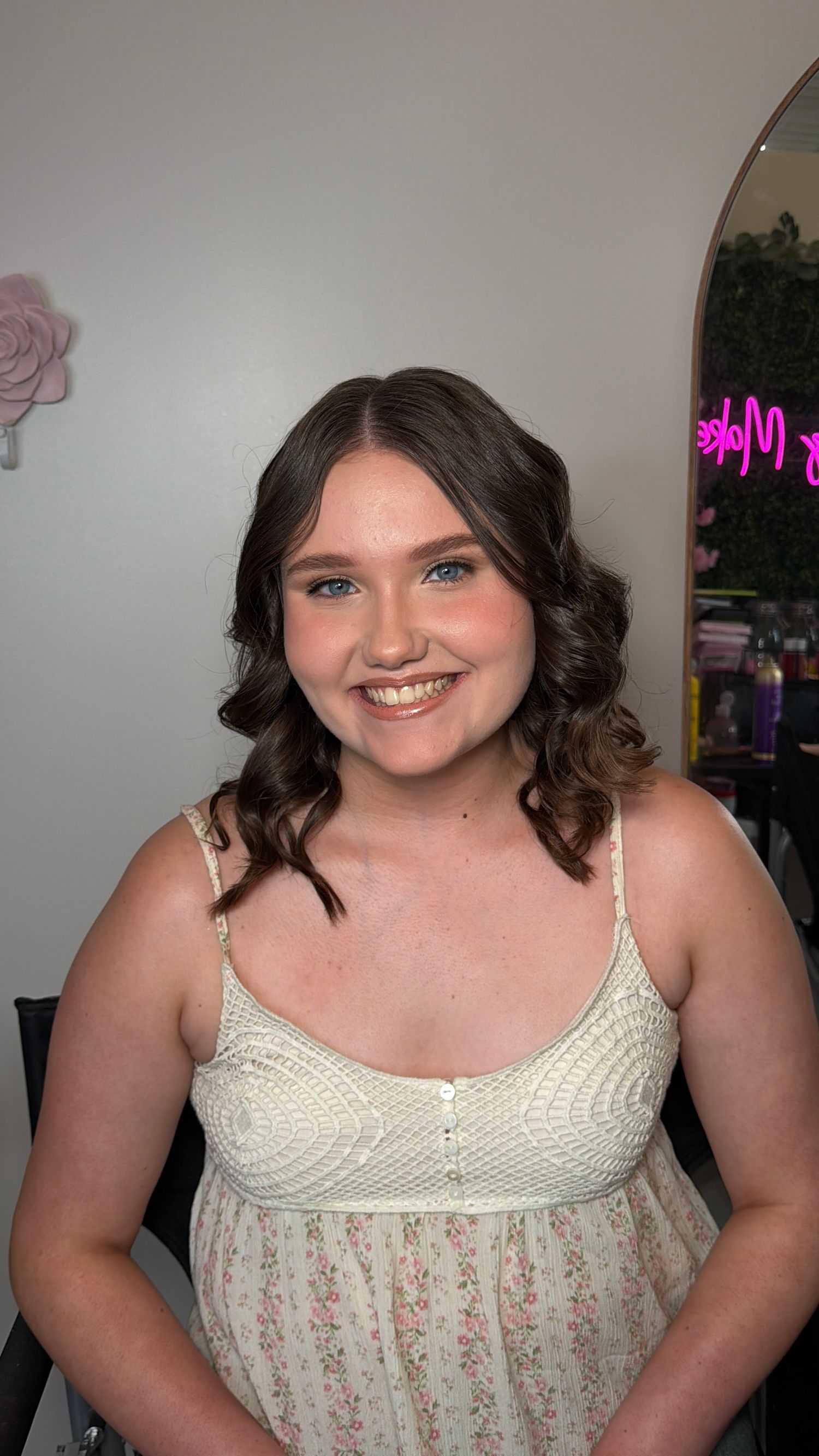 Smiling young woman with curly brown hair wearing a floral sundress in a beauty salon setting.