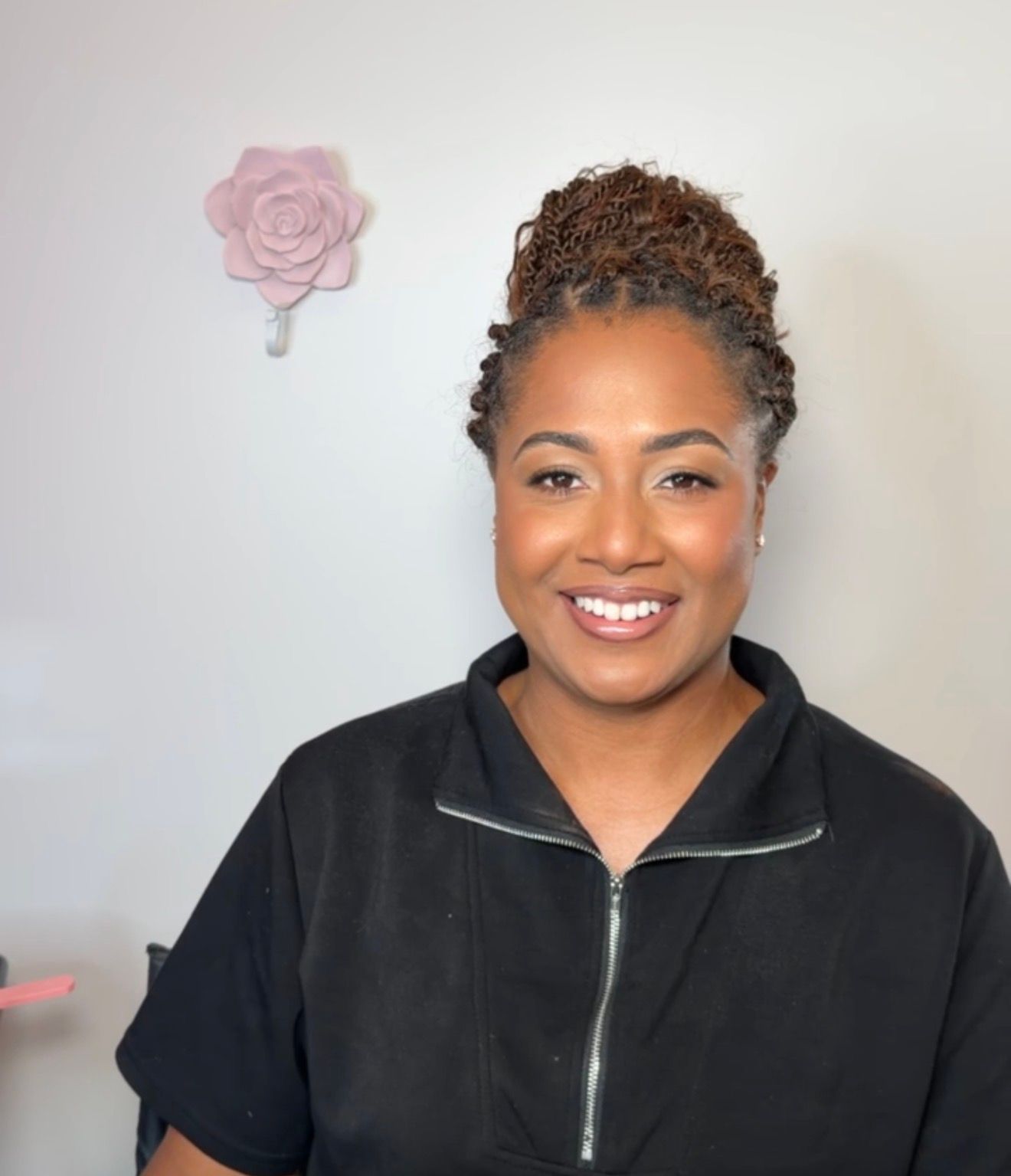 Smiling woman with curly updo hairstyle wearing black zip-up top, posing in front of white wall with pink rose decor.