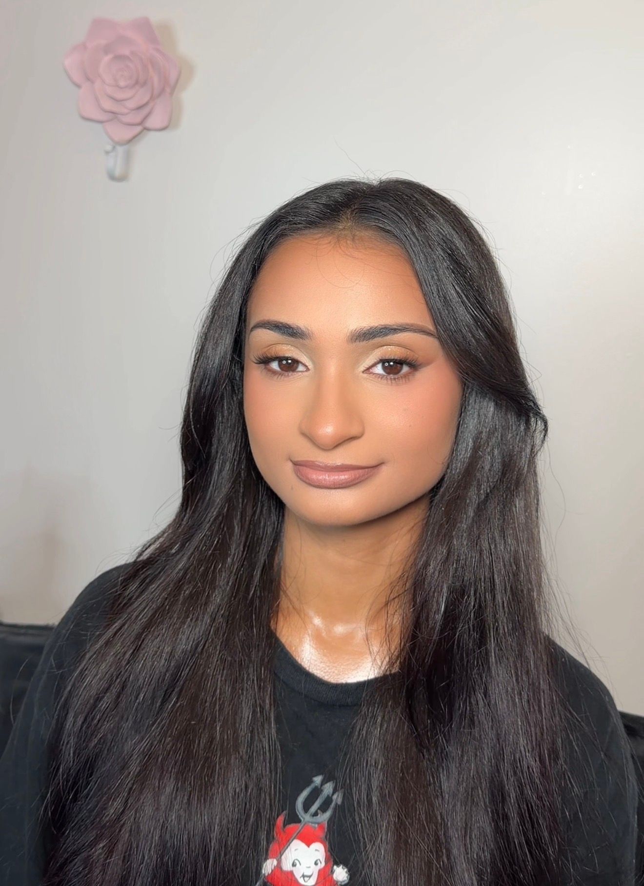 Young woman with long black hair and glam makeup wearing a black graphic tee, posing indoors near a pink rose decor.