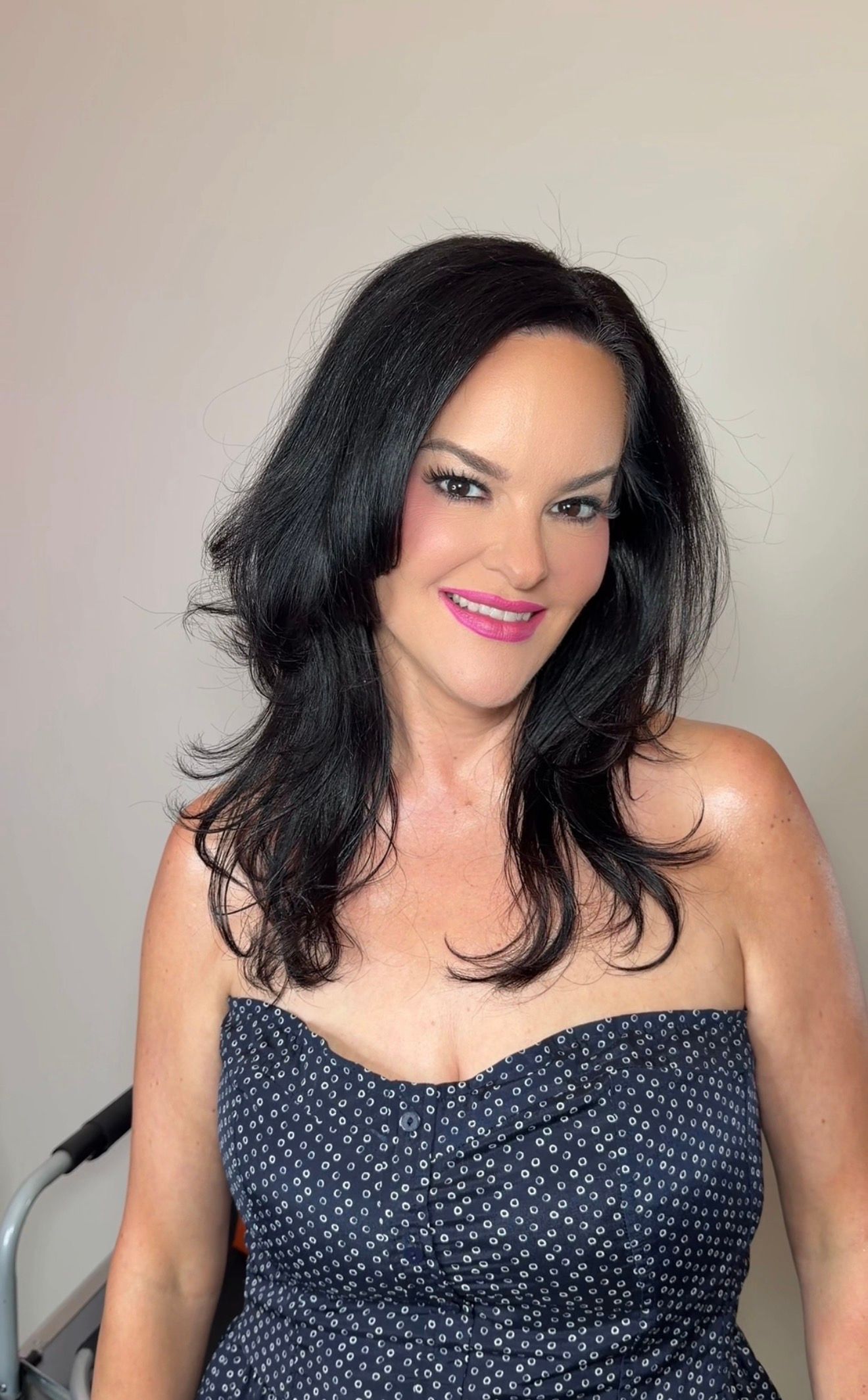 A woman with long black hair wearing a strapless navy polka dot dress, smiling with pink lips against a neutral background.