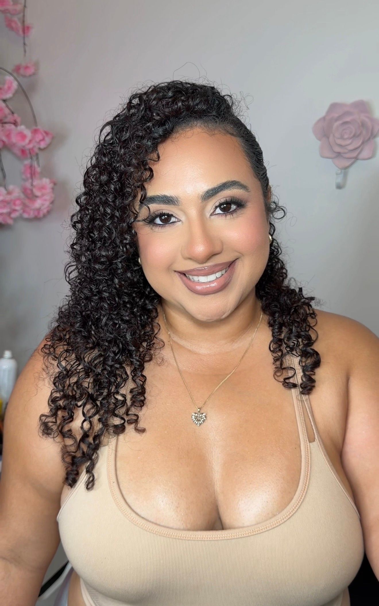 Smiling woman with curly hair wearing a beige top and heart necklace, posing in front of pink floral decor with glam makeup.