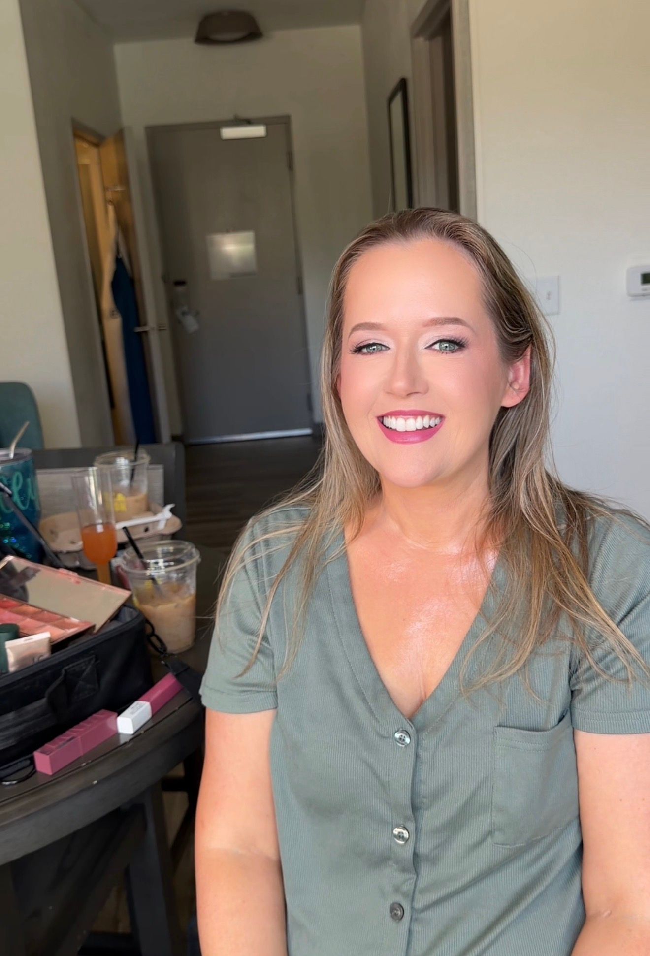 Smiling woman with long blonde hair wearing a sage green top, sitting near a makeup table in a modern room for her wedding day.