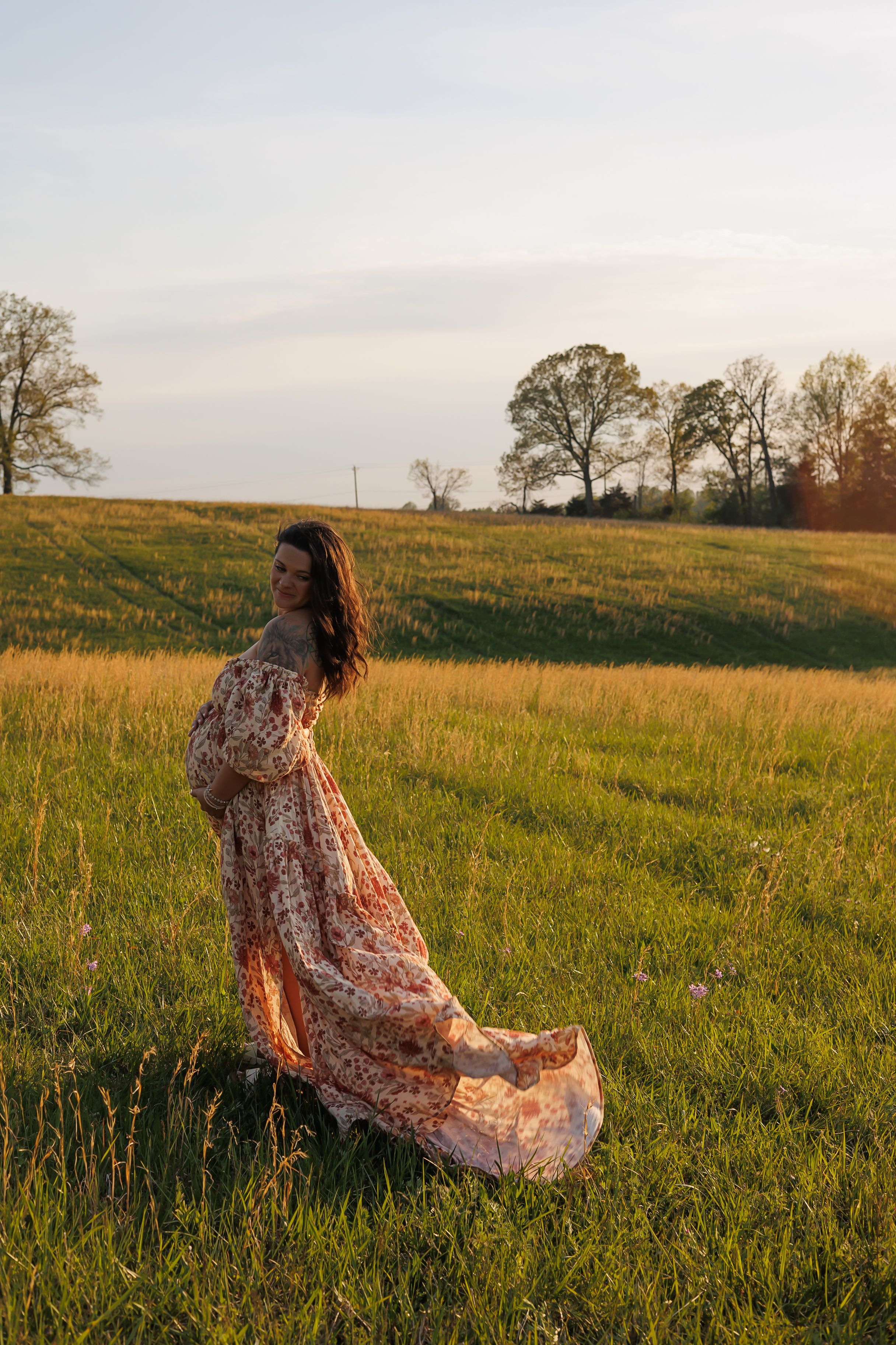 A flowy floral dress catches the golden sunlight in a serene meadow at sunset with trees in the background.