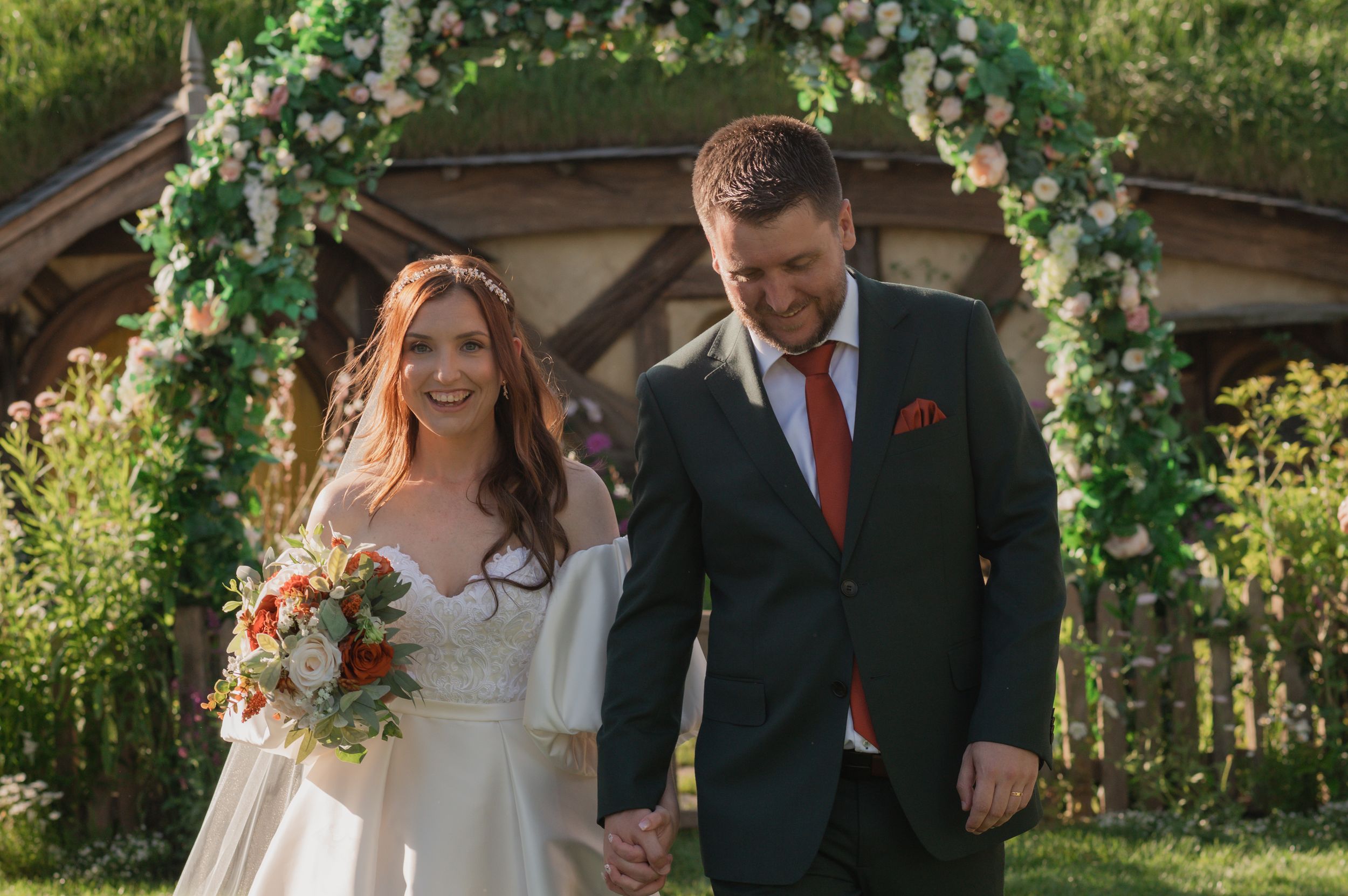 Bride and groom walk hand in hand under a floral arch outdoors on their wedding day.