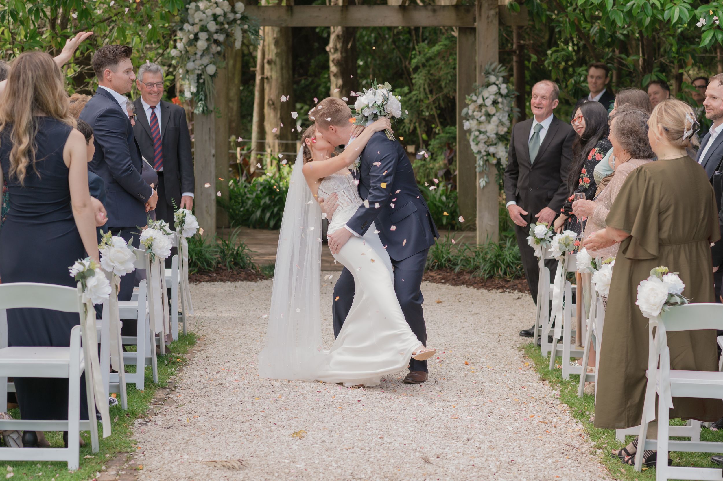 Bride and groom share a romantic kiss at outdoor wedding ceremony aisle lined with white chairs and floral arrangements.