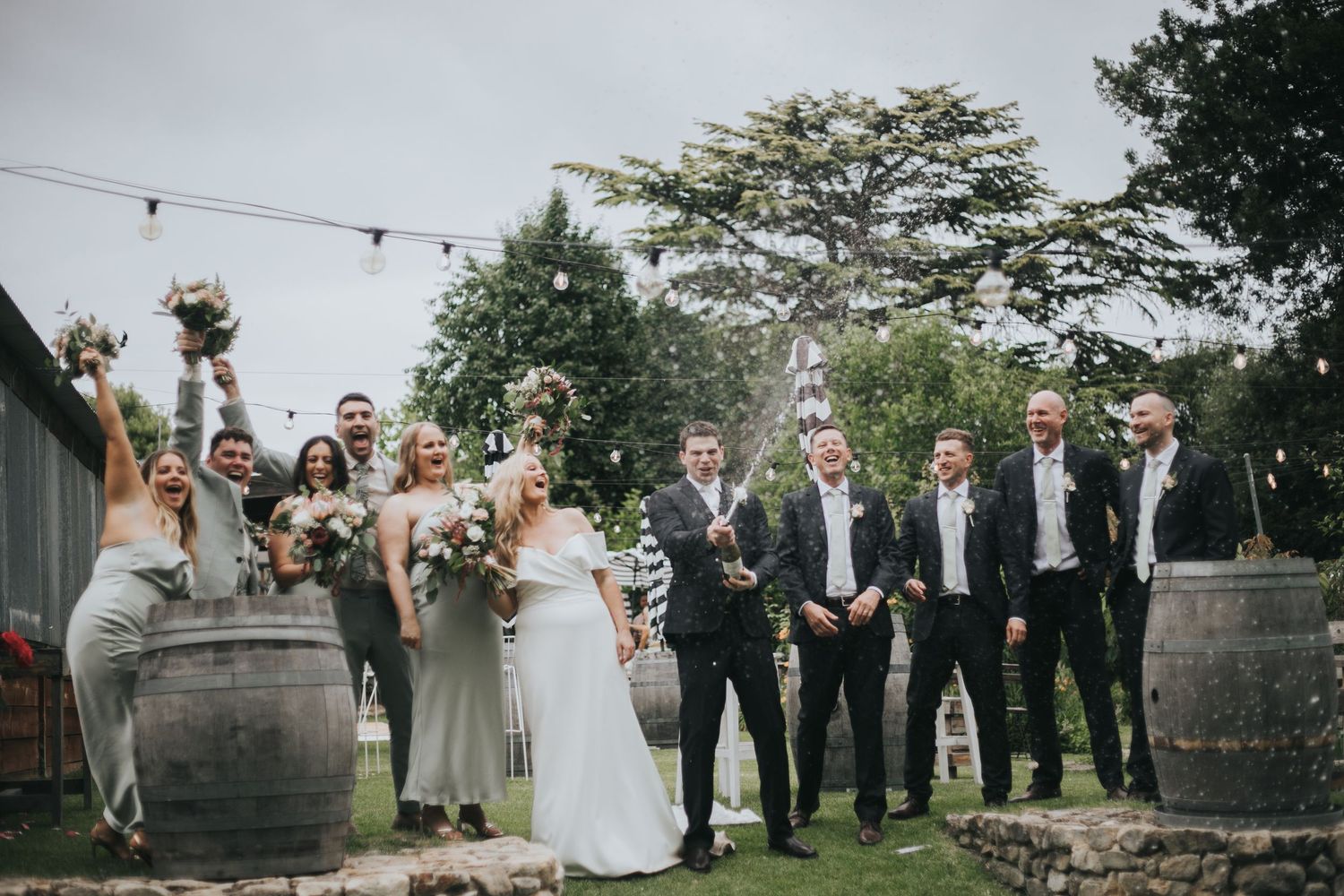 Wedding party celebrates with champagne beside wooden barrels at outdoor garden venue with festoon lights.