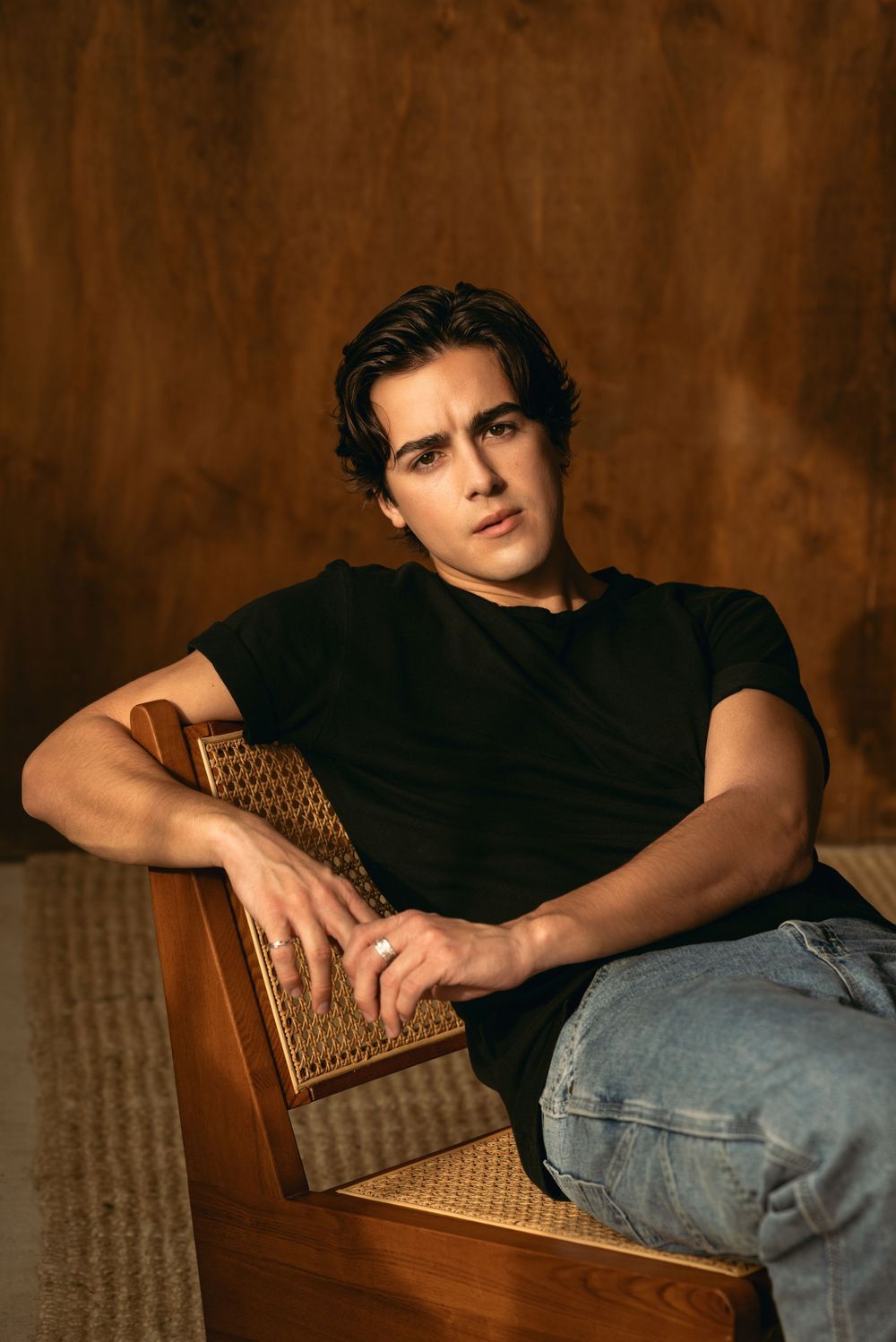 A person in a black shirt and blue jeans reclines on a textured brown chair in a moody studio setting.