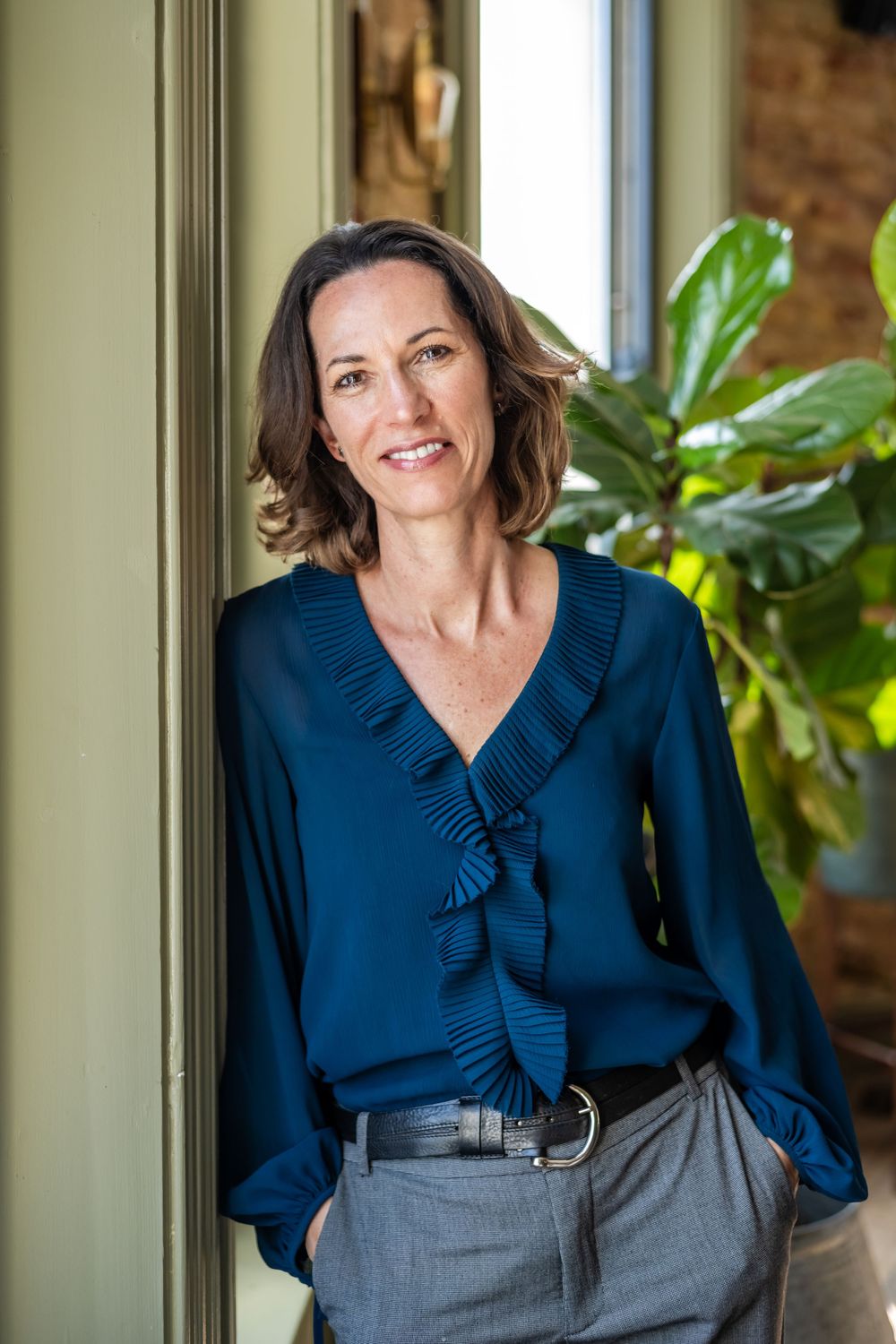 Professional headshot of woman wearing a navy blue blouse with pleated collar detail against a soft interior background.