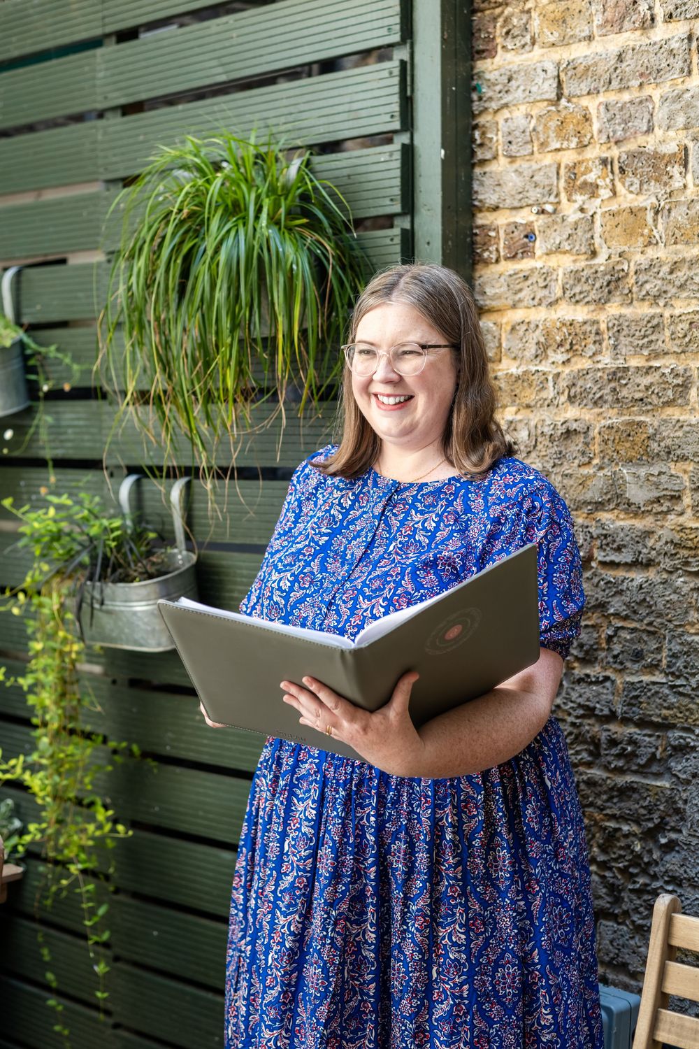 A person wearing a blue patterned dress stands by a brick wall with plants, holding an open folder and smiling.