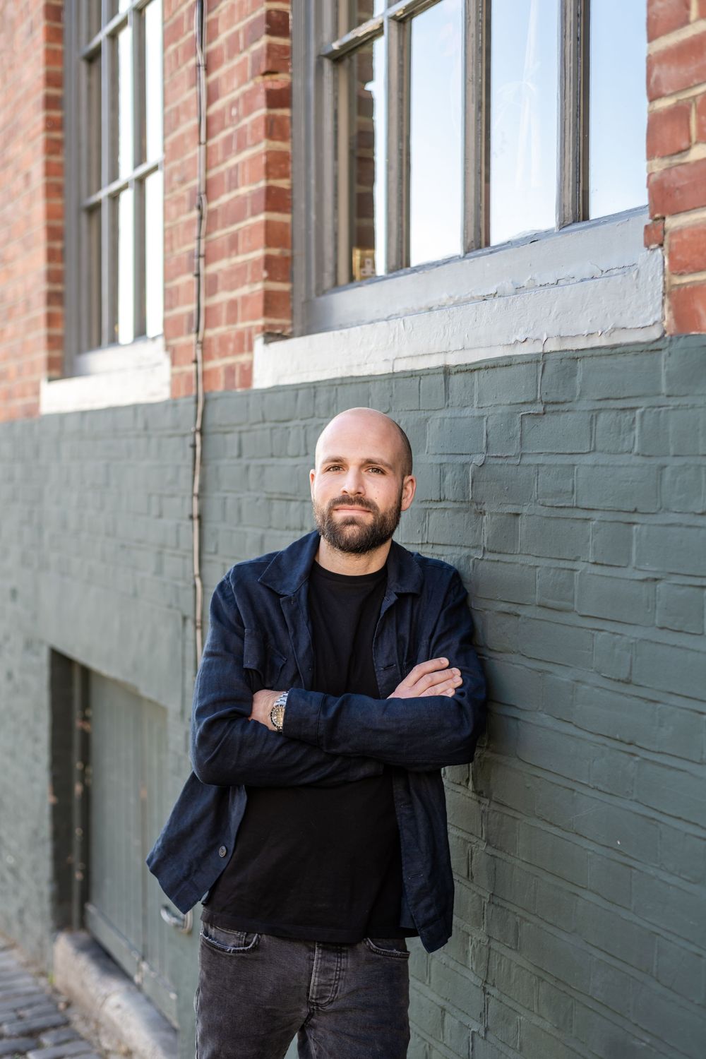Someone stands casually with arms crossed against a green painted brick wall wearing a dark jacket and jeans.