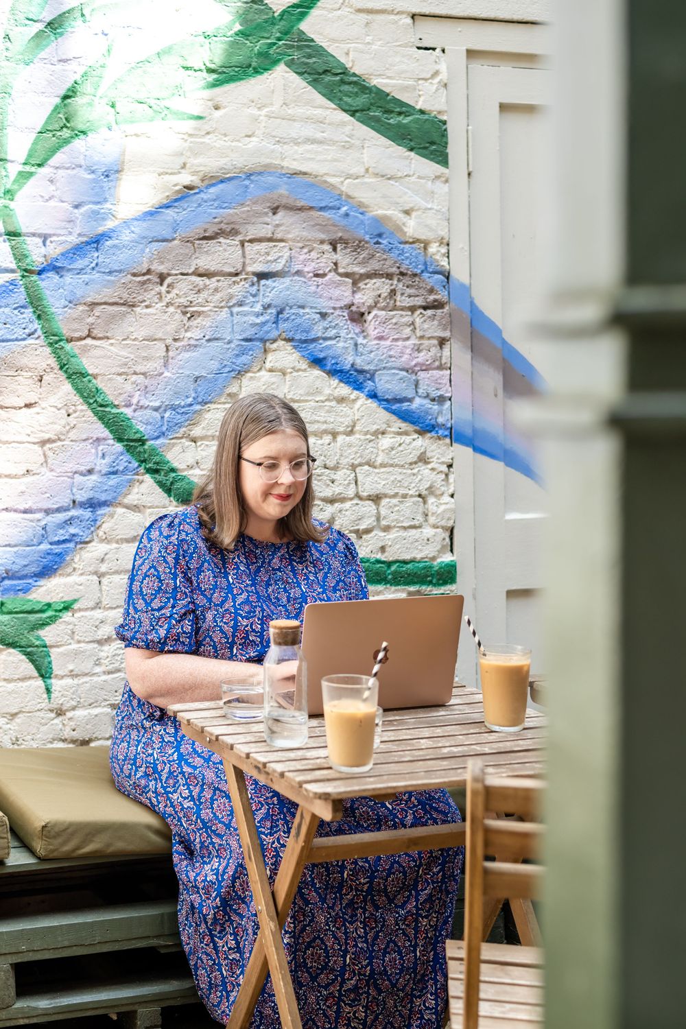 Someone in a blue patterned dress works on a laptop at an outdoor cafe table with iced coffee drinks next to a mural wall.