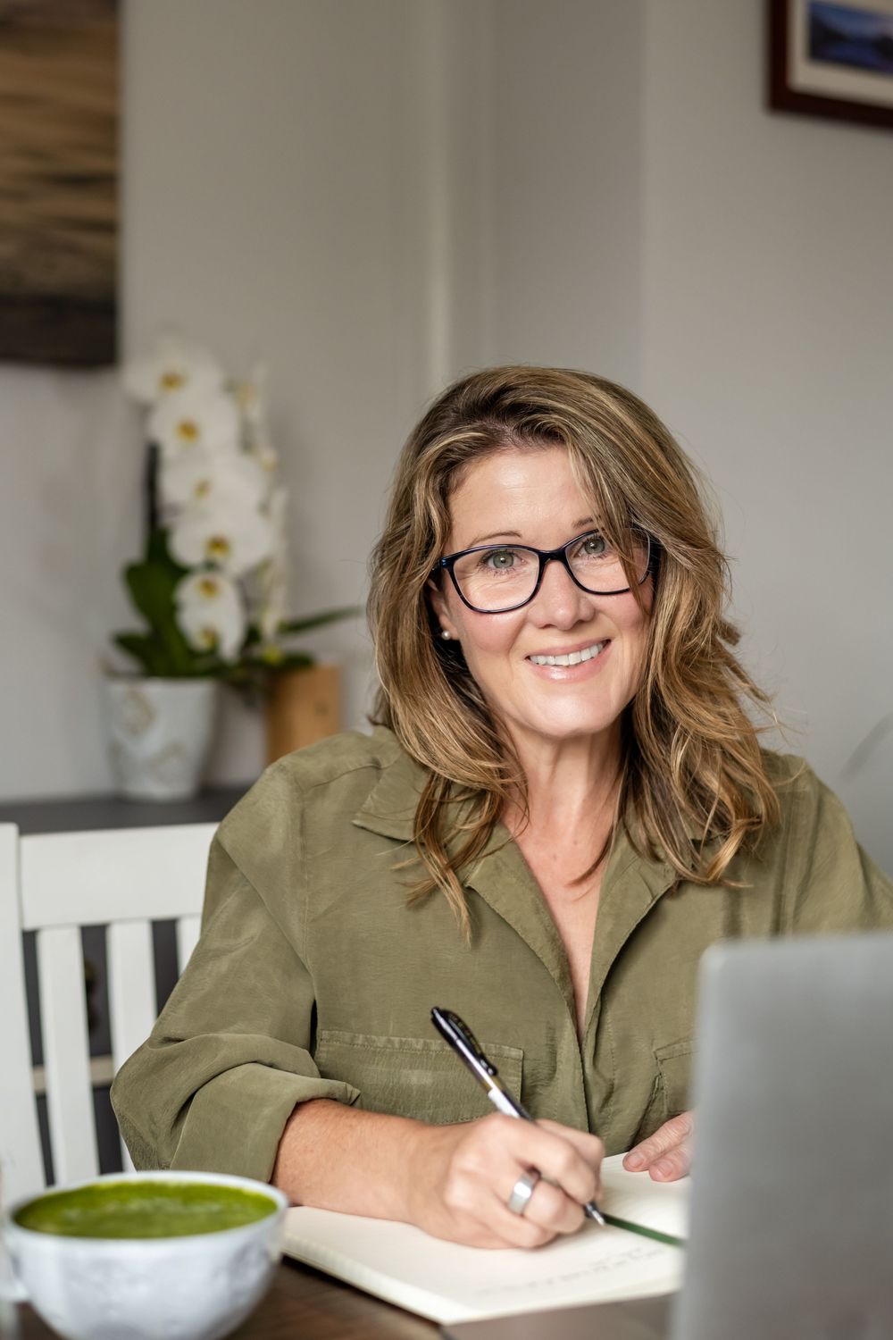Woman in glasses and olive shirt writes in notebook while enjoying green smoothie at white desk with orchid.