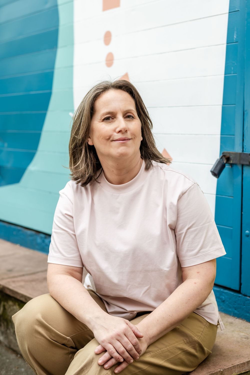 A person in a pink t-shirt and khaki pants sits casually against a blue and white wall backdrop.