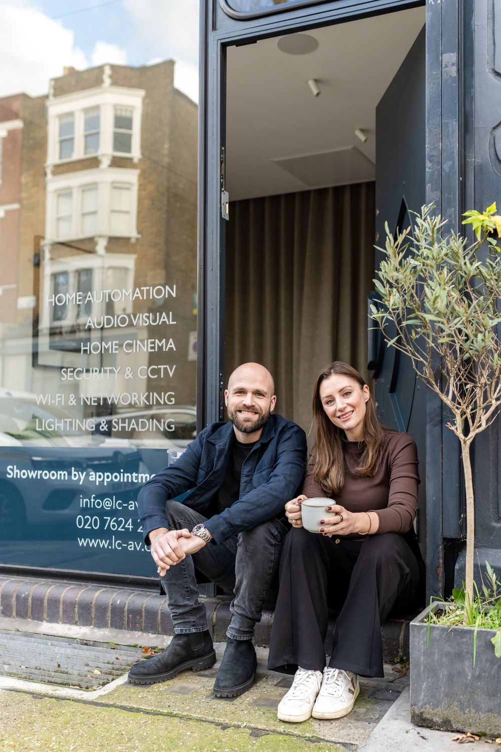 People sitting casually on a storefront step in front of a home cinema and lighting business window display.
