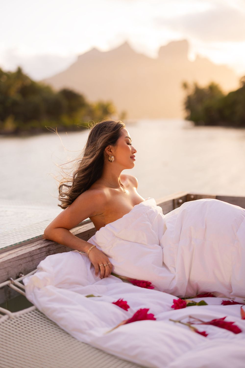 Romantic sunset scene on a luxury deck with white bedding and rose petals overlooking mountain silhouettes.
