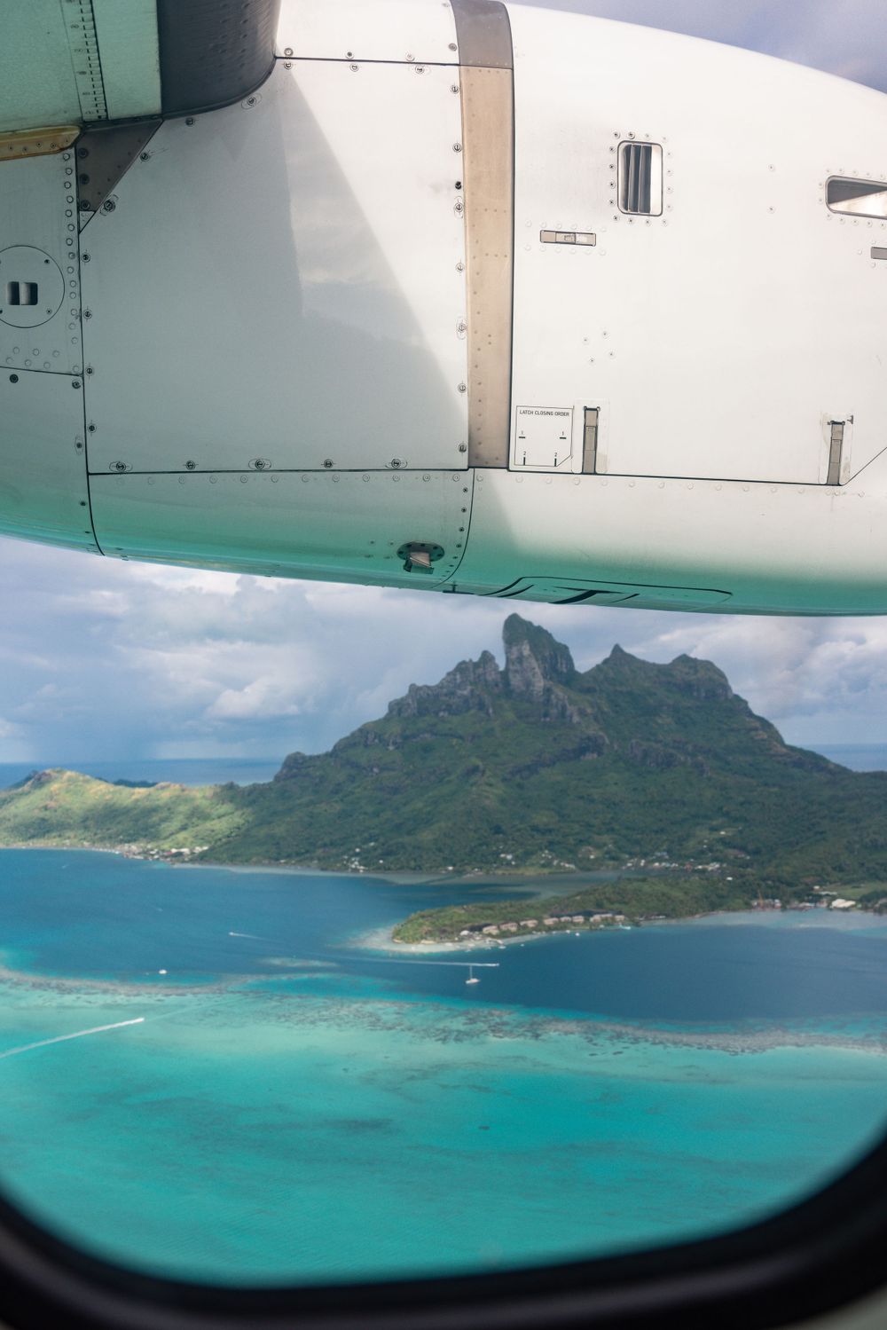 Aerial view of a tropical island with turquoise waters and dramatic mountain peaks seen through an airplane window.
