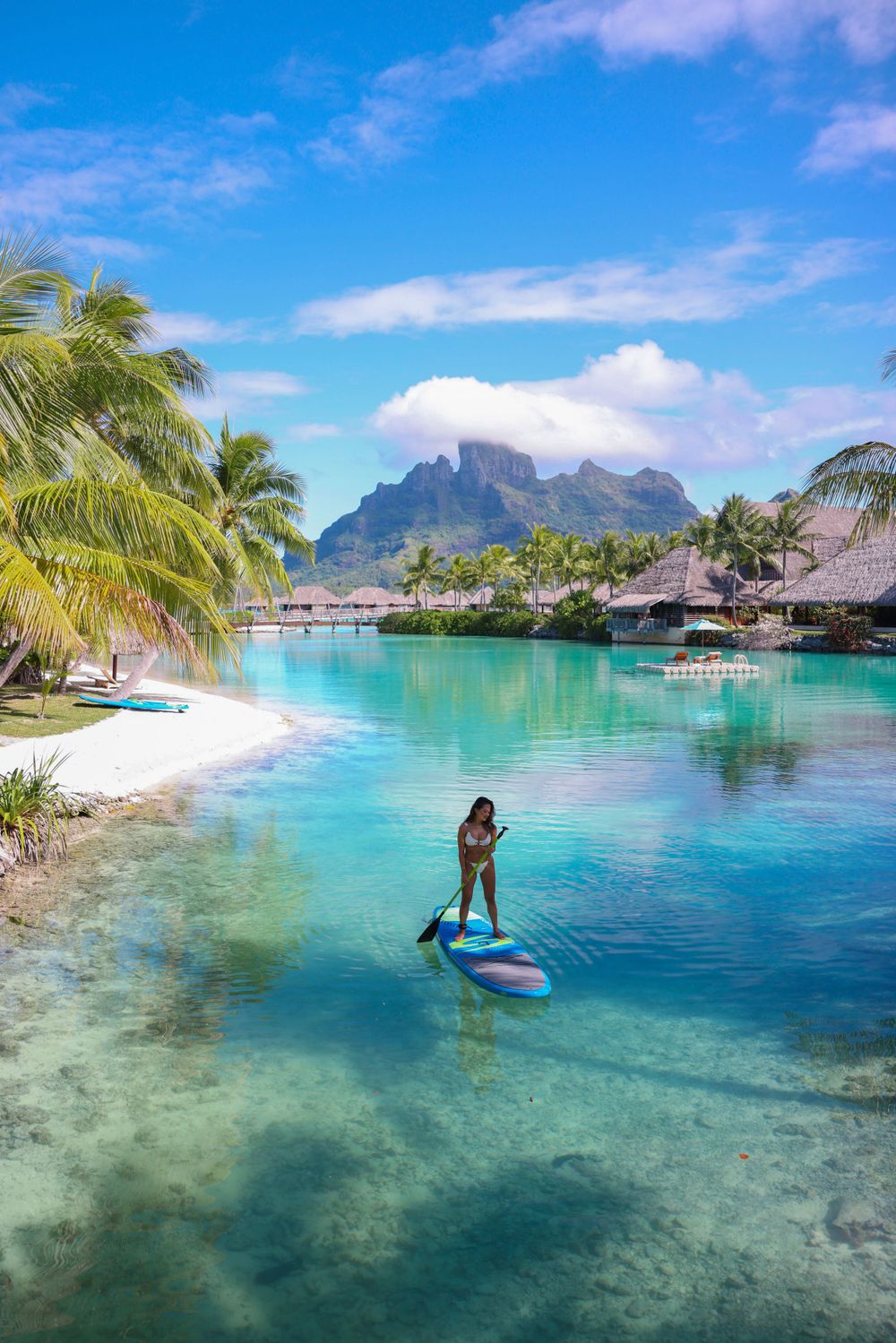 Paddleboarder on turquoise lagoon with overwater bungalows and mountain peaks of Bora Bora.