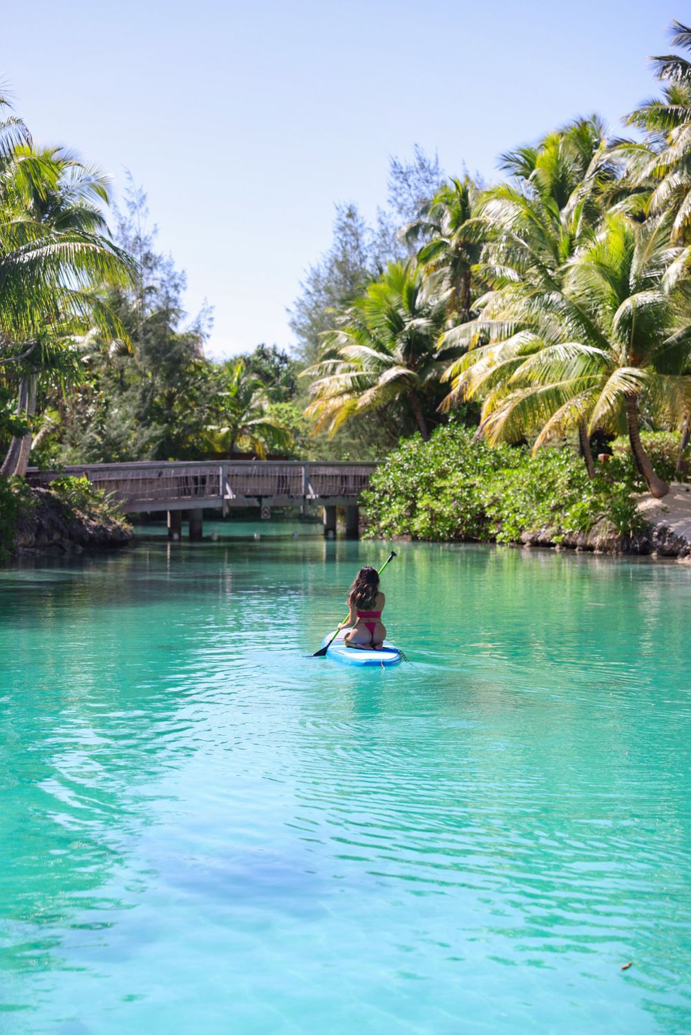 Paddleboarder gliding through crystal clear turquoise waters surrounded by palm trees and a wooden bridge.